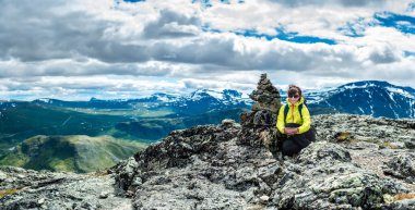 Jotunheimen, Norveç - 18 Temmuz 2016: Kız bir dağ manzarası ve bulutlu gökyüzünün arka planında bir taş yığınının yanındaki tepede oturuyor