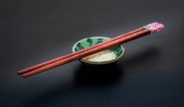 Yeşil bir kasenin tepesindeki ahşap yemek çubukları. Üzerinde pirinç ve koyu arkaplanda mavi harfler var..