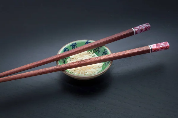 Yeşil bir kasenin tepesindeki ahşap yemek çubukları. Üzerinde pirinç ve koyu arkaplanda mavi harfler var..