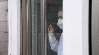 Maskeli adam, pencereden bakıyor. Koronavirüs için karantinada bir adam var..
