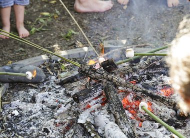 Çocuklar açık ateşte marşmelov pişiriyor. Serbest çocuklar, denetimli ve araştırmak için özgürler. Görüntü, NZ 'deki ateş çukurunda çocukların ayaklarını, lokumlu bambu çubuklarını, közleri ve küçük kütükleri gösteriyor. Çocuklarla kamp, ateş çukuru, ev okulu, vahşi..