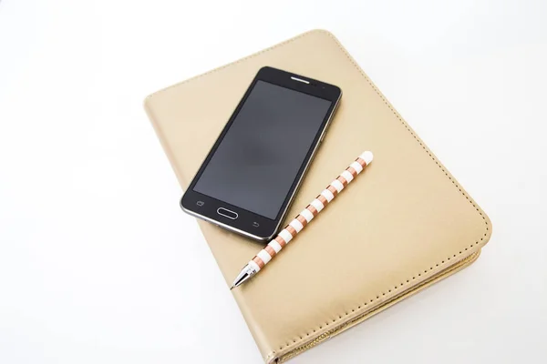 Black smart phone similar to leading brands sitting on top of gold compendium or diary with bronze and white striped pen. Useful for a contact me or communication page on a website or business. Working from home, home school, high school, university.