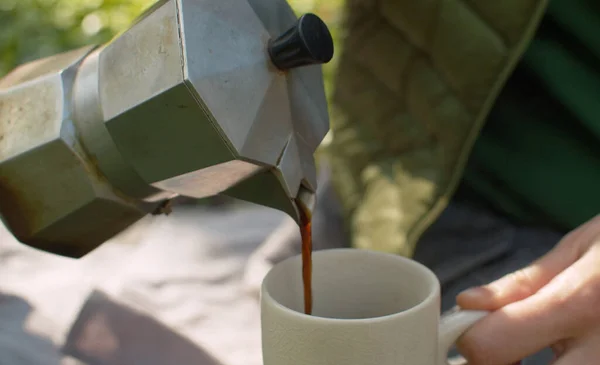 Close up female hand pouring coffee into a cup from the espresso maker. Steam comes out of it. People on picnic in the forest. Springtime, healthy lifestyle concept