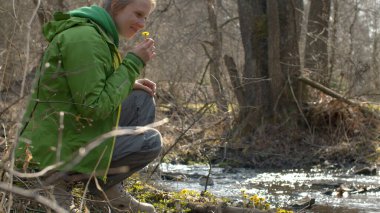 Nehir kıyısında oturan genç ve güzel bir kadın. İlk bahar çiçeğini seçiyor - tay ayağı. Nehirdeki su güneşte parlar. Çevre dostu kavram.