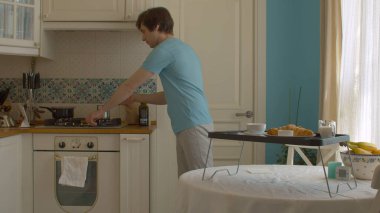 Genç adam mutfakta kahvaltı hazırlıyor. Cemve 'i yangından alıyor. Kruvasan tabağı, reçelli fincan ve tepside sütle dolu kavanoz var..