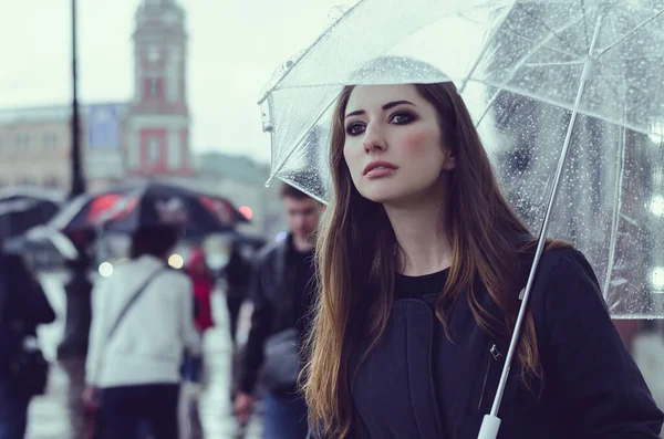 Film grain image with pretty young woman in a coat holding a transparent umbrella, standing in a crowd on rainy day in a city center. Movie screenshot in cold colors. Accessory or lifestyle concept