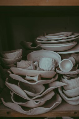 Raftaki çeşitli kurumuş kil sofra takımlarının yakın çekimi. Çömlek stüdyosu konsepti. El yapımı tabaklar, kaseler, tencereler, farklı renkte kupalar. Samimi bir manzara. Seramik bulaşık takımı ile atölye.