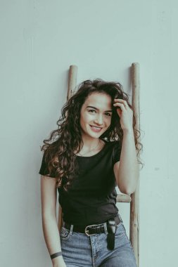 Siyah tişörtlü ve kot pantolonlu gülümseyen Avrupalı bir kadın beyaz duvarın önünde duruyor ve arka planda merdivenler var. Mutlu bir çömlekçi kadın. Seramik stüdyo konsepti