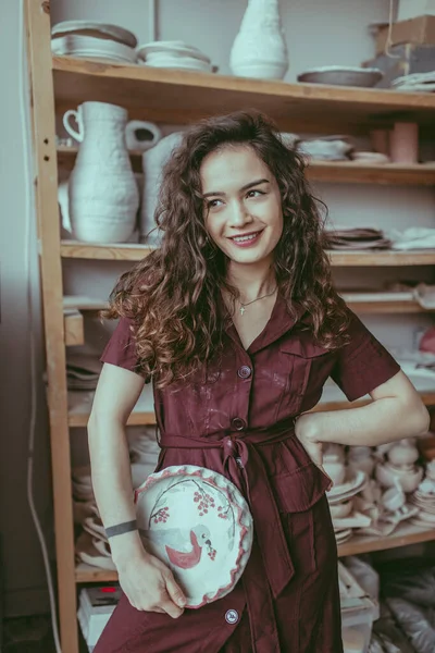 A smiling european young pottery woman with long curly hair holding a handmade dish in her hand. A ceramist in her ceramics studio at work. A girl in red dress stained with white paint