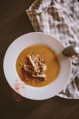 Balkabağı kremalı çorba ve kremalı beyaz tabak, ayçekirdeği ve kraker çorbaya eklendi. Üstü kırmızı biberle kaplı. Tabağın yanında doğal renklerden oluşan keten bir havlu ve bir kaşık dolusu gümüş var..