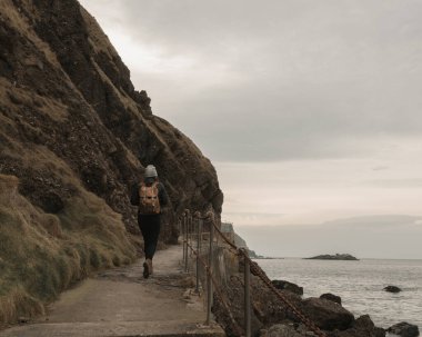 Tanımlanamayan, şapkalı ve sırt çantalı bir kız denizin kenarında yürüyor.