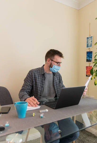 Side view of young european man wearing a face-mask working from home due to the corona-virus restrictions with a laptop and papers