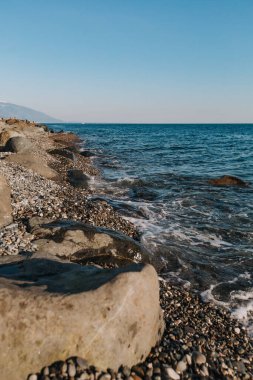 Mavi deniz, masmavi gökyüzüne ve uzaktaki dağlara karşı kiremitli plajı ve büyük kayaları yıkıyor..