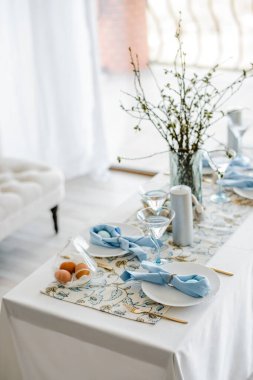 Gökyüzü mavisi tekstil peçetesi ve çiçek masa örtüsüyle dekoratif masa örtüsü, paskalya yumurtaları, tabaklar ve altın çatal bıçak takımı, bardaklar. Vazoda söğüt ağacı. yazlık ev içi.