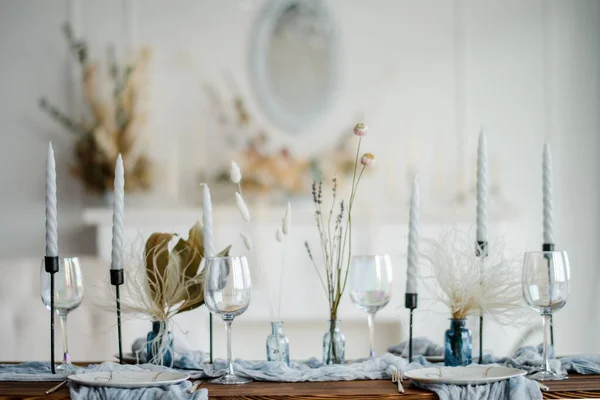 Stylish table setting with dried flowers. Plate with vintage golden fork and knife, candles, dusty blue napkins on wooden table. Winter wedding decoration.