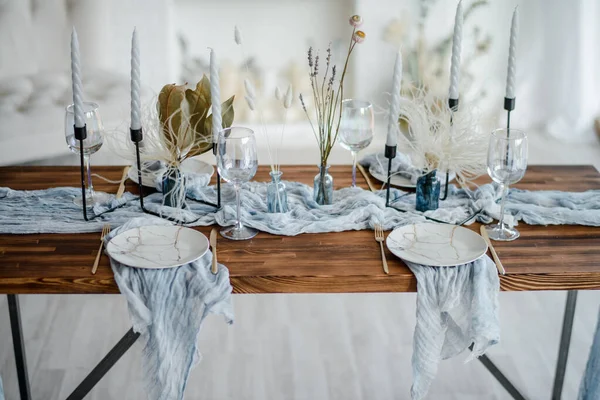 Romantic table setting for holiday dinner, wooden table served with dried flower, plates, golden cutlery, white candeles, bright dusty blue runner. Selective focus.