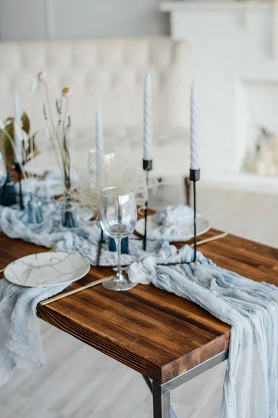 Romantic table setting for holiday dinner, wooden table served with dried flower, plates, golden cutlery, white candeles, bright dusty blue runner. Selective focus.