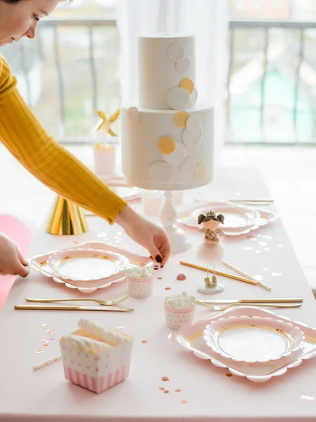 Woman serving party table with cake in pink and white colors, gentle textile tablecloth, paper dishes, cups and golden cutlery. Happy birthday party for little girl.