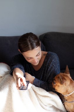 Kanepede oturan genç ve güzel bir kadın yanında Fransız buldoguyla televizyon seyrediyor.
