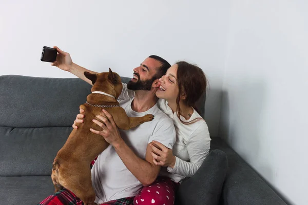 Genç bir çift köpekle birlikte evdeki kanepede selfie çekiyor.