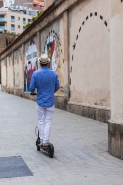 Şapkalı genç adam elektrikli scooterıyla şehri geziyor.