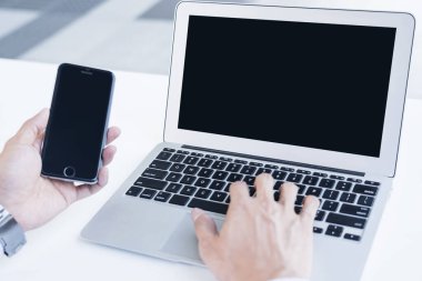 Adam dizüstü bilgisayar kullanıyor ve elinde akıllı telefon tutuyor.