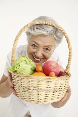 Yaşlı kadın elinde sebze ve meyve dolu bir sepet tutuyor.