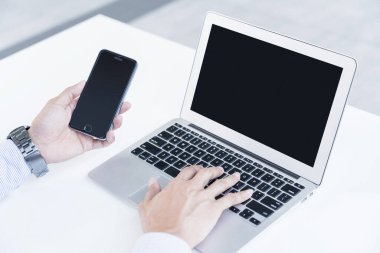 Adam dizüstü bilgisayar kullanıyor ve elinde akıllı telefon tutuyor.