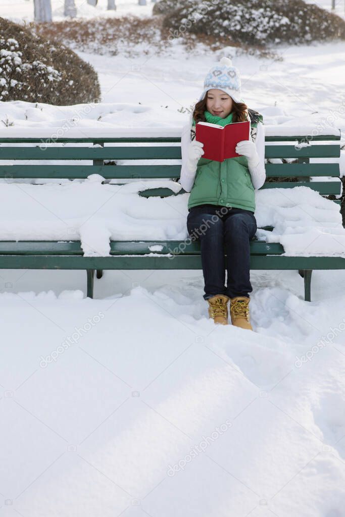 Woman in warm clothing reading book on a snowy bench