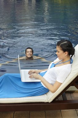 Havuz başında dizüstü bilgisayar kullanan bir kadın, havuzda yüzen bir adam.