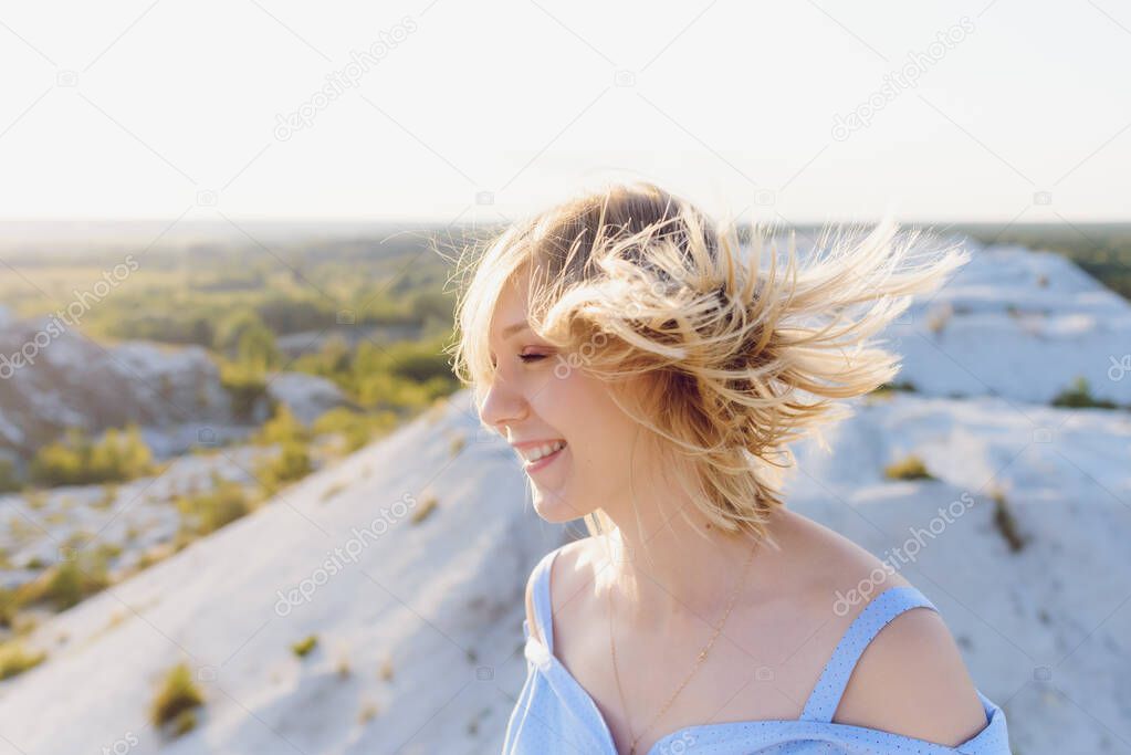 Portrait of smiling beautiful girl outdoors. Young girl standing on a mountain with white hair covering her face when blown by the wind