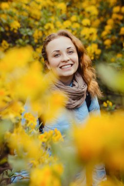 Sarı rhododendronların ve büyük yaprakların arka planında dalgalı saçları olan mavi ceketli güzel bir kız. Moda, güzellik, sağlık, kişisel bakım, doğa.