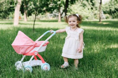 Small baby girl in dress walking on grass and rolling toy stroller with doll in park on summer clear day. Happy childhood concept clipart