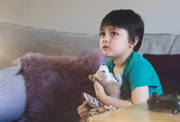 Emotional portrait of kid sitting on sofa hugging his soft toy looking up with worrying face,Child with sad face while watching cartoon on TV, Little boy stay at home relaxing in living room