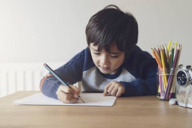 Masada oturup ödev yapan okul çocuğunun portresi, elinde kalem tutan mutlu çocuk masadaki beyaz kağıda resim çizen bir çocuk, ilkokul ve evde eğitim kavramı.