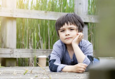 Outdoor portrait of happy boy lying on wooden bridge and looking at camera with curious face,Active child having fun playing in wildlife park,Kid having adventure in nature reserve in sunny day summer clipart