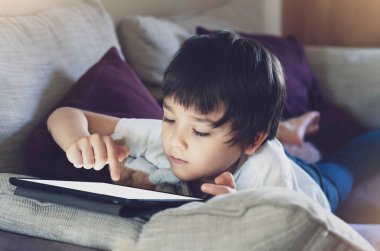Ödevi için tablet kullanan çocuk izolasyonu, kanepede uzanmış çizgi film seyreden ya da dijital tablette oyun oynayan çocuk, evde eğitim, sosyal uzaklık, e-öğrenim online eğitim.