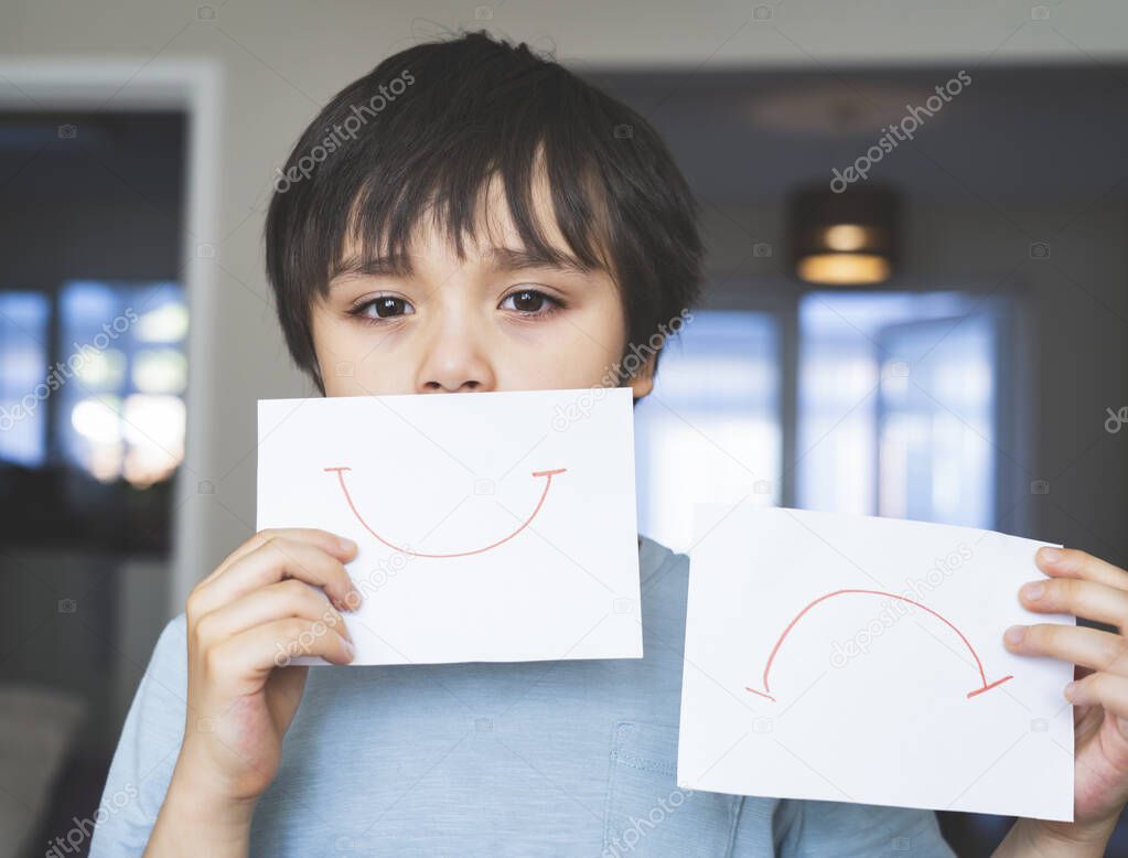 Portrait of bored kid with sad face holding  white paper with smile and sad,  Child boy getting bore stay at home during during self-isolation, quarantine. Coronavirus outbreak and flu covid epidemic 