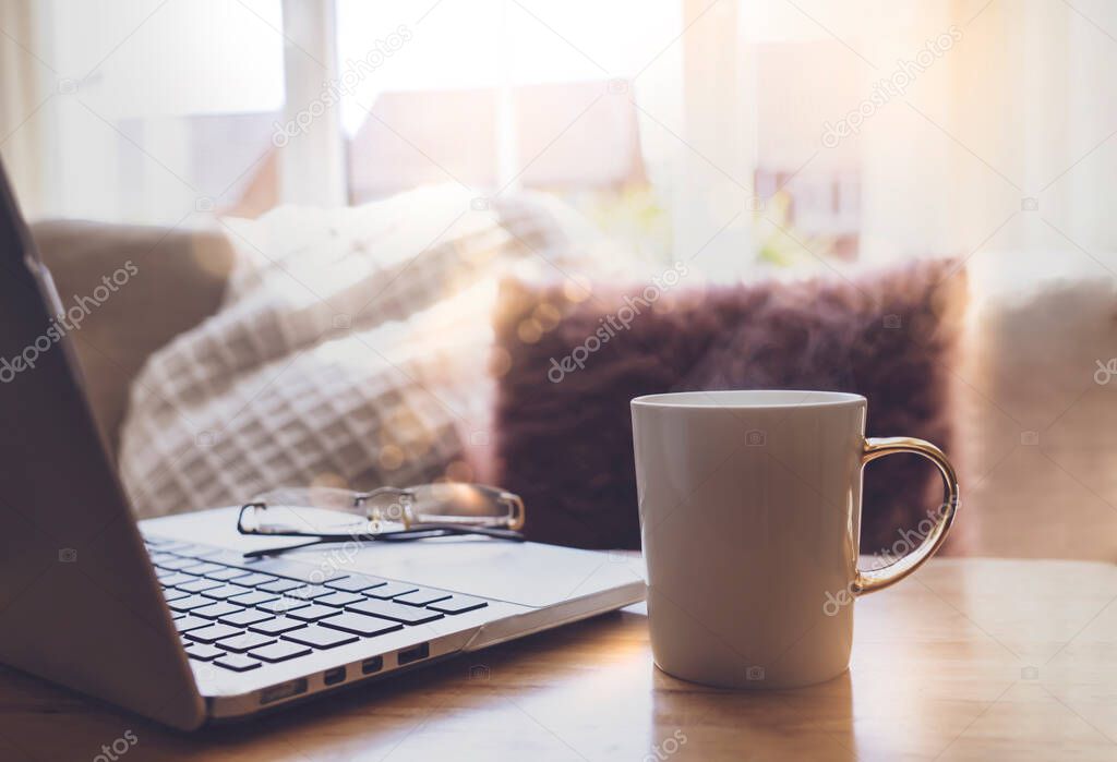 Soft focus mug of aroma tea with laptop and blurred cushion background next to window in the morning or Cozy Work space in afternoon tea with notebook and hot coffee,New in normal life at home concept