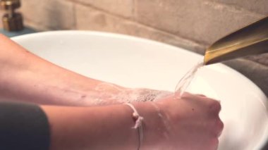 Beyaz oval bir lavabo kâsesinde ellerini sabunla yıkayan ve durulayan genç bir kadın.