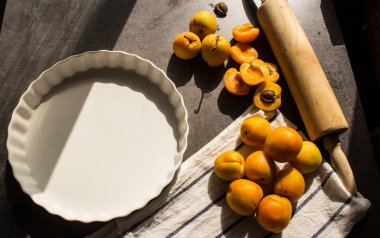 Kayısılı kek yapıyorum. Beyaz pişirme formu, hamur rulosu ve mutfak masasında taze meyve. Koyu gri arkaplan. 