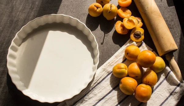 Kayısılı kek yapıyorum. Beyaz pişirme formu, hamur rulosu ve mutfak masasında taze meyve. Koyu gri arkaplan. 