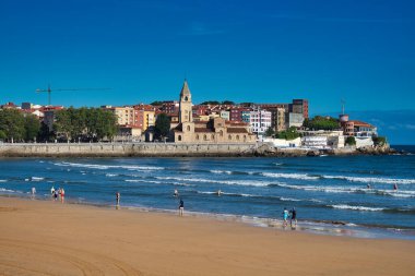 San Lorenzo plajı, sahilde yürüyen insanlar, denizde dalgalar ve önünde San Pedro kilisesi olan komşular, Gijon, Asturias, İspanya.