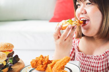 Asyalı şişman bir kadın evindeki kanepede pizza ve kızarmış tavuk yiyor. Hastalığa neden olan gıda tüketimi konsepti sağlık için iyi değildir.