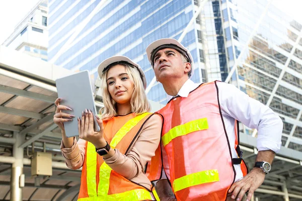 Western male and female engineers wear white safety hats and carry tablets. Standing for meetings outside the building Point your finger to the top to check the job.