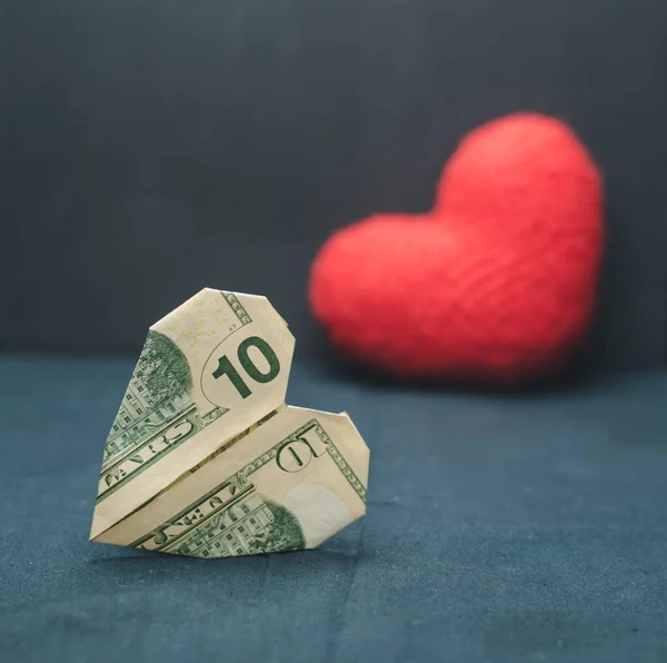 A heart made of paper money in the foreground and a red heart in the background, dark background. Romance. Mothers Day. Valentine\'s Day.