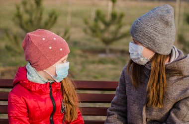 Tıbbi maskeli kızıyla birlikte bir bankta oturan bir anne. Coronavirus koruyucu maskesi. Covid-19. Tıbbi maskeli çocuklu genç bir kız.