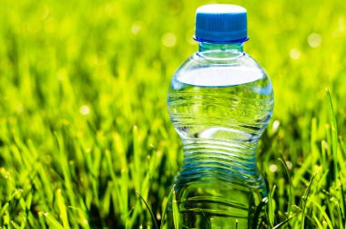 Su şişesi çimlerde. Çimlerin üzerinde güneş ışınlarıyla şeffaf plastik su şişesi. Güneş ışınları su şişesinde parlıyor..