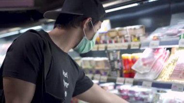 Cerrahi maske takan genç Asyalı markete alışverişe gidiyor, yiyecek stokluyor, rafın önünde duruyor, alışveriş sepeti tutuyor, kovid19 Corona virüsü salgını sırasında yaşam tarzı, 4k