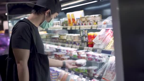若いアジアの男性は 医療用マスクに黒いキャップを着用します食料品店で買い物に行きます 食品の在庫 棚の前に立って 健康的な食品の選択 Covid 19コロナウイルスパンデミックの間のライフスタイル — ストック動画